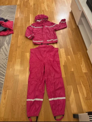 Rosa regnkläder med reflexdetaljer - Regnkläder med avtagbar huva och reflexband över bröst, rygg och ärmar. Perfekt för blöta dagar. Spännbara hängslen. I superbra skick förutom att bandet vid ena foten är lite slitet, se sista bilden. Från ICA 