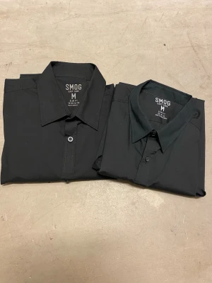 2 St Svarta Skjortor, Långärmade  - 2 Skjortor från Smog i storlek M. Köpta på New Yorker för 150kr styck och har inte använts, dvs inga defekter som fläckar, hål, osv. Mitt pris är 100kr för båda två. Dvs 50kr/styck, så du sparar 66% / 200kr.         Frakt sker inom 24, skriv vid frågor 