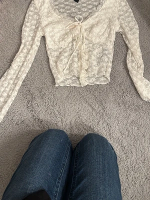 Vit spetsblus med knyt och volang - Säljer en superfin vit blus i spets med långa ärmar. Toppen har knyt framtill och en söt volangdetalj längs knäppningen. Den är croppad och har en romantisk vibe, perfekt att styla med jeans eller kjol.