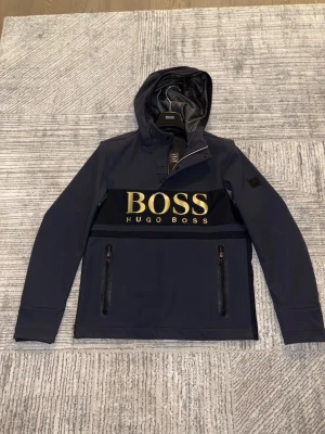 Hugo Boss jacka - Säljer en marinblå vindjacka från Hugo Boss med stor logga i guld på bröstet. Jackan har huva, dragkedjor på fickorna och detaljer i svart. Materialet är lätt och vattenavvisande syntet, perfekt för blåsiga dagar. Snygg och sportig look med modern passform.