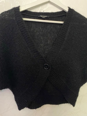 Svart stickad bolero med fladdermusärmar - Kort svart stickad kofta med v-ringning och en stor knapp framtill. Koftan har bred ribbad kant och är perfekt att slänga över en topp för en chill look. Passar dig som gillar oversized och mysiga plagg. Den är glittrig i färgen.