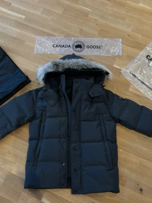 Grå pufferjacka från Canada Goose - Säljer en grå pufferjacka från Canada Goose med huva och snygg fuskpäls runt kanten. Jackan har klassisk patch på ärmen, dragkedja och knappar framtill. Perfekt för kalla dagar och riktigt stilren look.