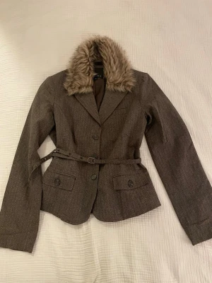 Brun jacka med päls - Brun jacka/kavaj med diskreta ränder. Jackan har en avtagbar pälskrage och ett bälte. Märke Amisu, storlek 38. mått: från armhåla till armhåla 45 cm, jacklängd 58 cm, ärmlängd från utsidan 62 cm, från insidan 48 cm.