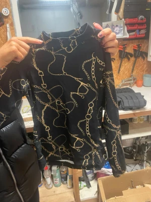 Svart långärmad topp från CRW - Snygg svart långärmad topp från CRW med guldfärgade kedjemönster över hela plagget. Hög hals och figurnära passform, perfekt för dig som gillar en trendig och lite lyxig stil. Materialet känns mjukt och följsamt.