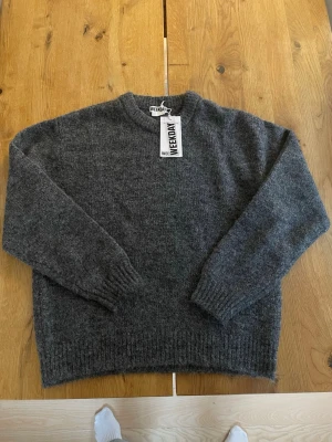 Mörkgrå stickad tröja från Weekday - Mysig mörkgrå stickad tröja från Weekday i storlek S. Aldrig använd Tröjan har en klassisk rund halsringning, ribbade muddar vid ärmslut och nederkant samt lång ärm. Perfekt för kyliga dagar och enkel att styla med olika outfits. 