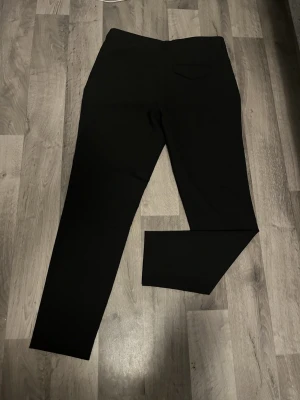 Svarta kostymbyxor från Arken - Säljer ett par klassiska svarta kostymbyxor från Arken. Byxorna har rak passform, diskret bakficka med lock och stängs med knapp och dragkedja. Perfekta till skjorta eller stickad tröja. Materialet känns mjukt och följsamt.