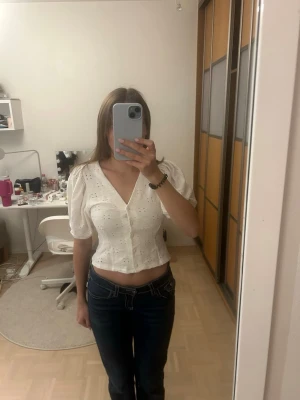 Vit blus med puffärm från H&M - Säljer en söt vit blus från H&M i storlek XS. Blusen har puffärmar, v-ringning och knappar framtill. Materialet är bomull med broderade detaljer och små hålmönster. Croppad passform som ger en fräsch och somrig vibe.