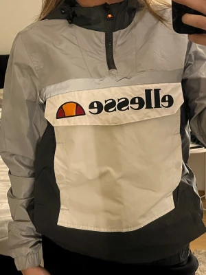Grå och svart vindjacka från Ellesse - Säljer en snygg vindjacka från Ellesse i grått och svart med stor logga framtill. Jackan har huva med snörning, halv dragkedja på sidan för tajtare passform och stor magficka samt fickor på sidan. Den sitter tajtare i midjan och skapar en attraktiv passform på kroppen. 