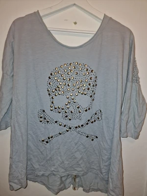 Ljusblå topp med nitar och spets - Unik ljusblå topp med trekvartsärm, dekorerad med nitar i döskallemönster framtill och spetsdetalj bak. Har en avslappnad oversize vibe om man i vanliga fall har en mindre storlek. 