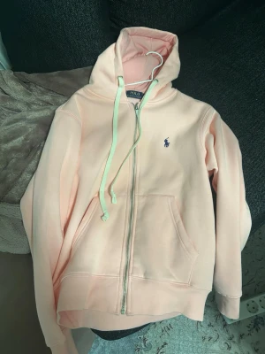 Ralph Lauren zip rosa  -   	Ljusrosa zip-hoodie från Polo Ralph Lauren i storlek S. Mjuk och bekväm med fleece på insidan, dragkedja framtill och broderad logga på bröstet. I fint skick, sparsamt använd. 