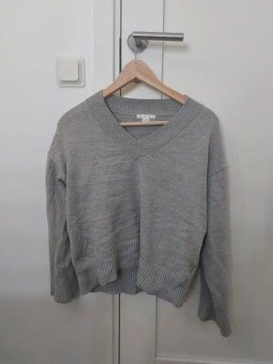 Grå stickad tröja från Gina Tricot - Mysig grå stickad tröja från Gina Tricot med bred ribbad v-ringning, muddar vid ärmslut och nederkant. Perfekt för lager på lager och chill dagar. Passar dig som gillar en enkel men snygg stil.