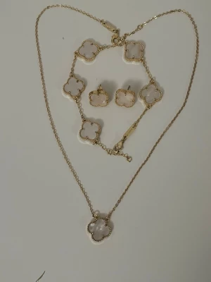 Fyrklöver smycken set 18k guld - Säljer ett snyggt smyckesset med halsband, armband, ring och örhängen i vit och guld. Alla delar har fyrklöverformade hängen med guldkant. Haösband, armband, örhängen och ring. Perfekt för dig som gillar stilrena accessoarer med en touch av lyx.