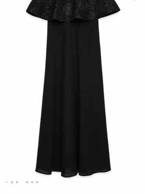 Svart axelbandslös långklänning - Säljer en svart axelbandslös långklänning med volang upptill och diskret blommönster på överdelen. Helt ny med prislappen kvar