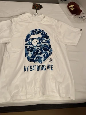 Vit BAPE t-shirt med blå camo print - Säljer en clean vit t-shirt från BAPE med det ikoniska apansiktet i blått camo-mönster på bröstet och texten 'BY BATHING APE' under. Klassisk passform och crewneck. Perfekt för dig som gillar streetwear och vill sticka ut. Storlek s men passar m 