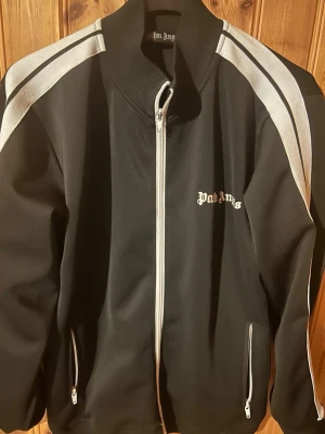 Palm Angels svart track jacket - Säljer en svart track jacket från Palm Angels med vita ränder längs ärmarna och vit dragkedja. Tröjan har hög krage, två fickor med dragkedja och logga på bröstet. Perfekt för en sportig och trendig look.