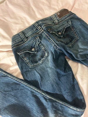 Blå jeansbyxor med broderade fickor - Säljer ett par blå jeansbyxor från Rock & Republic i storlek W31/L32. Jeansen har snygga broderade bakfickor med dekorativa knappar och kontrastsömmar. Klassisk femficksmodell med normal passform. Jag säljer jeansen då de är för stora för mig 