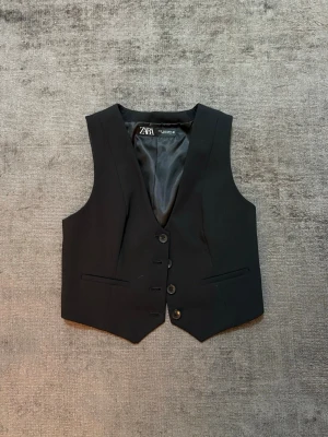 Svart kostymväst från Zara - Säljer en svart kostymväst från Zara med klassisk v-ringning och knäppning framtill. Västen har två dekorativa fickor och är fodrad med slätt tyg. Perfekt att styla över en skjorta eller t-shirt för en trendig look.