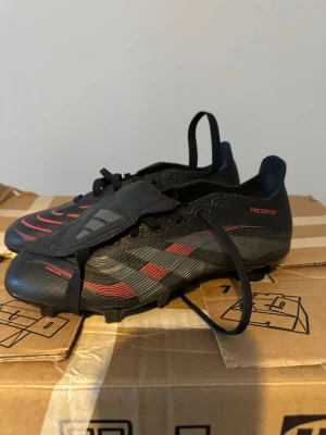 Adidas Predator fotbollsskor svart/röd - Säljer ett par Adidas Predator fotbollsskor i svart med röda och grå detaljer. Skorna har snörning, täckt plös och mönstrad ovandel för extra grepp. Perfekta för dig som vill dominera på planen med stil.
