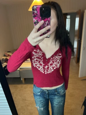 Röd broderad topp från D&G - Säljer en röd långärmad topp från D&G med vit broderad design framtill. Toppen har v-ringning med knappar och är croppad i modellen. Perfekt för dig som gillar unika detaljer och vill sticka ut lite extra.