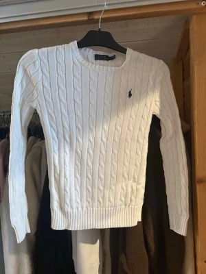 Vit kabelstickad tröja Polo Ralph Lauren - Kabelstickad vit tröja från Polo Ralph Lauren i klassisk modell med rund hals och lång ärm. Tröjan har det ikoniska mörkblå logobroderiet på bröstet och ribbade muddar vid ärmslut och nederkant. Perfekt till jeans eller kjol för en clean look.