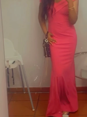 Rosa offshoulder långklänning - Säljer en snygg rosa långklänning med offshoulder-design och v-ringad omlottdetalj upptill. Klänningen har en figurnära passform och en liten slits nertill för extra rörelsefrihet. Perfekt för dig som vill sticka ut med en färgstark look.