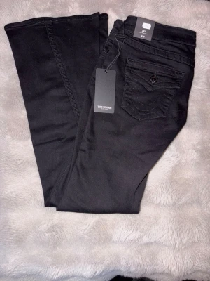 Svarta True Religion Joey flare jeans - Säljer ett par svarta True Religion Joey jeans med låg midja och flare-ben. Byxorna har snygga detaljerade bakfickor med knapp och dekorativa sömmar. Klassisk femficksmodell i mjukt bomullsmaterial med stretch för extra komfort.