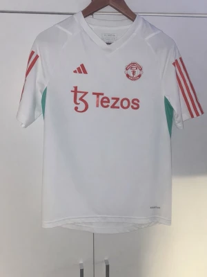 Manchester United adidas tröja 13-14Y - Säljer en vit Manchester United matchtröja från adidas med röda detaljer och gröna inslag på sidorna. Tröjan har korta ärmar, Tezos-logga på bröstet och klassiska adidas-ränder på axlarna. Perfekt för dig som älskar fotboll och vill ha en snygg supportertröja.