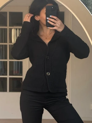 Svart stickad kofta med knappar Stockh LM - Säljer en svart stickad kofta med snygga detaljer och två stora knappar framtill. Koftan har lång ärm, bred krage och ribbade muddar. Perfekt att slänga på sig när det är kyligt ute.