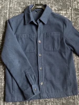 Mörkblå overshirt i ull - Säljer en stilren mörkblå overshirt från Donce de la sense i mjuk ullmix. Jackan har klassisk krage, två bröstfickor och knäppning framtill med stora knappar. Perfekt lager-på-lager-plagg med enkel, clean look och lång ärm.
