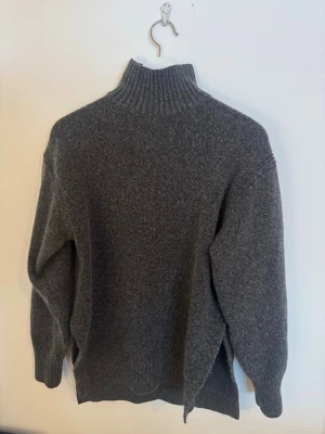 Grå polotröja i ull med slits - Mysig mörkgrå polotröja i ull med ribbad hög krage och slits i sidorna. Tröjan har lång ärm och en loose fit som ger en chill vibe. Perfekt för kalla dagar när du vill vara både varm och snygg.