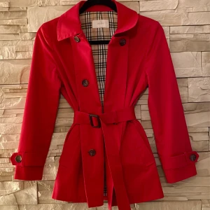 Röd trenchcoat från Zooc med bälte - Snygg röd trenchcoat från Zooc med klassiskt rutigt foder. Jackan har knytskärp i midjan, dubbelknäppning framtill och detaljerade knappar vid ärmsluten. Långärmad modell med fickor och tidlös krage. Perfekt för dig som vill sticka ut i höst eller vår.