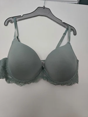 Grön/Grå spets-bh från Redcent 85C - Snygg hrön/grå bh från Redcent i storlek 85C. Bh:n har vadderade kupor och fina spetsdetaljer längs nederkanten och sidorna. Axelbanden är justerbara och knäppningen bak är klassisk med hyskor. Materialet är mjukt och stretchigt i polyester och elastan.