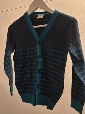 Randig blå kofta med knappar - Snygg randig kofta i mörkblått och turkost från Polarn o Pyret. Koftan har långa ärmar, v-ringning och knäppning framtill med fyra knappar. 