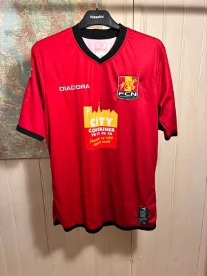 FC Nordsjælland Diadora 2013/15 shirt, storlek M - Ok skick.  #fcnordsjælland #danmark #superliga #john #hansen #vingaard #runje  Röd FCN matchtröja med sponsorlogotyper. Tröjan har svarta detaljer vid halsringningen och ärmsluten. Klubbmärket och Diadora-logotypen är placerade på bröstet.