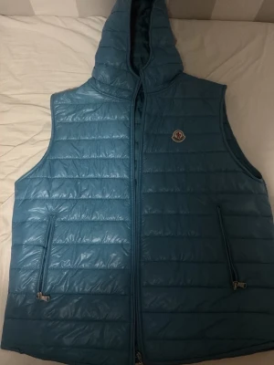 Blå dunväst med huva från Moncler - Säljer en blå quiltad dunväst från Moncler med huva och dragkedja framtill. Västen har två fickor med dragkedja och Moncler-logga på bröstet. Perfekt för lager-på-lager och riktigt snygg till streetwear-stilen.