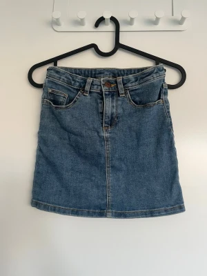 Blå jeanskjol med fickor - Säljer en klassisk blå jeanskjol i kort modell med fem fickor och bälteshällor. Kjolen har en rak passform och stängs med knapp och dragkedja framtill. Perfekt att matcha med sneakers eller boots för en avslappnad look.