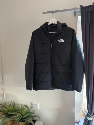 Äkta Svart pufferjacka från The North Face - Säljer en svart pufferjacka från The North Face med huva och dragkedja framtill. Jackan har en stilren design med diskret logga på bröstet. Perfekt för kalla dagar och enkel att matcha med allt. Pris går att diskutera!🙌