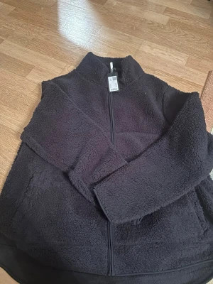 Svart teddyjacka från Lager 157 - Mysig svart teddyjacka från Lager 157 . Jackan har hög krage, dragkedja framtill och två sidofickor. Perfekt för kalla dagar när du vill vara både varm och stilren. Materialet är fluffigt och ger en riktigt chill vibe. Något tunnare modell 