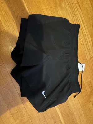 Svarta träningsshorts från Nike - Säljer ett par svarta träningsshorts från Nike med elastisk midja och snörning. Shortsen har en klassisk loose fit och är tillverkade i lätt polyester som andas. Perfekta för löpning eller gymmet. Nike-logga i vitt på ena benet. Innerbxor 