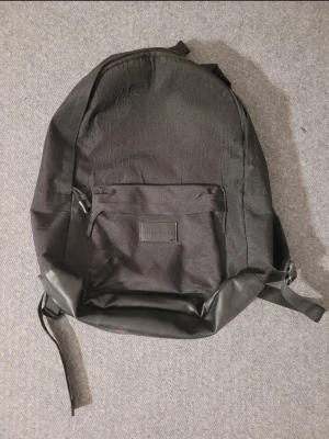 Svart ryggsäck från Pier One - Säljer en stilren svart ryggsäck från Pier One med en stor ytterficka framtill och detaljer i skinnimitation. Perfekt för att bära laptop, böcker eller gymkläder. Justerbara axelband och dragkedja upptill gör den smidig att använda.