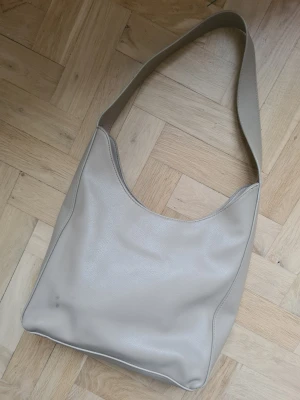 Beige axelväska från Åhléns - Beige axelväska med bred rem och rund form. Väskan har ett stort huvudfack och innerfack med dragkedja. Mindre skador, se bilder.