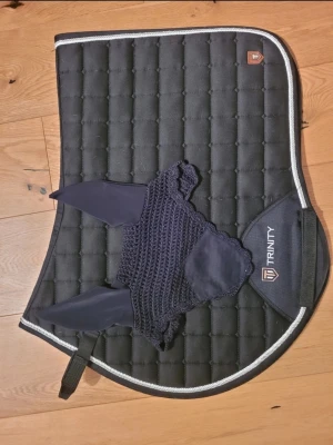 Svart schabrak från Trinity - Säljer ett svart, quiltat schabrak från Trinity med vit kant och matchande öronhuva i mörkblått. Perfekt för ridning och ger en clean look till din hästutrustning. Schabraket har en diskret logga och praktiska fästen.