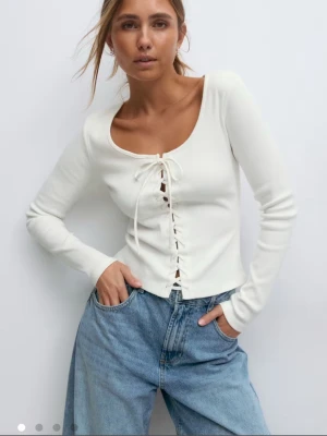 Vit ribbad topp med snörning - Säljer en vit långärmad topp med snygg snörning framtill och rundad urringning. Toppen är ribbad och har en tight passform. Helt oanvänd och prislapp kvar.