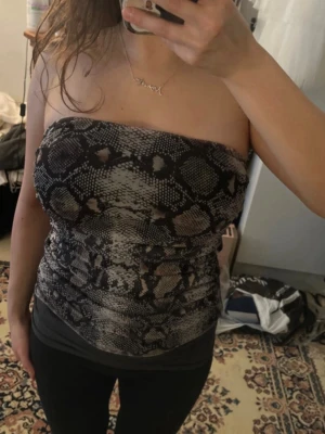 Tubtopp med ormmönster - Säljer en snygg tubtopp med ormmönster i svart och grått. Toppen är åtsittande och axelbandslös, perfekt för dig som vill sticka ut lite extra. Materialet känns mjukt och stretchigt, vilket gör den bekväm att bära hela dagen.