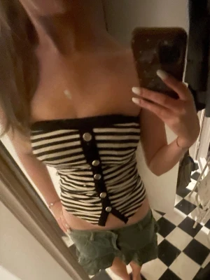 Randig bandeau tubtopp med knappar - Säljer en svartvit randig bandeau tubtopp med dekorativa silverfärgade knappar framtill. Toppen är ribbad och har en croppad passform. Perfekt för sommaren och enkel att styla till olika looks! Inbyggd bh och jätte bekväm!