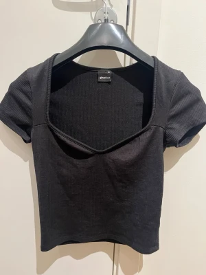 Svart ribbad topp från Gina Tricot - Säljer en svart ribbad topp från Gina Tricot i storlek XS. Toppen har korta ärmar, croppad passform och en snygg v-ringad urringning. Perfekt att matcha med höga jeans eller kjol för en trendig look.