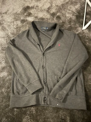 Grå kofta från Polo Ralph Lauren M - Mysig grå kofta från Polo Ralph Lauren i storlek M. Klassisk design med knappar framtill, två fickor och rosa broderad logga på bröstet. Tillverkad i mjuk bomull och perfekt för lager på lager under höst och vinter.