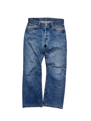 Levi's 501 blå jeans straight fit - Klassiska blå Levi's 501 jeans med rak passform och knappgylf. Jeansen har slitningar och hål på ena benet för en cool vintagekänsla. Tillverkade i tålig denim av bomull och har den ikoniska läderpatchen bak i midjan.