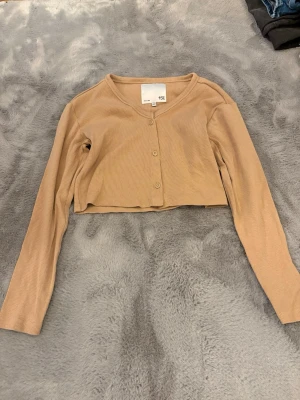 Beige croppad kofta från lager 157 - Säljer en beige croppad kofta från 157 med ribbad struktur och långa ärmar. Koftan har knappar framtill och en enkel, clean look som passar perfekt till lager på lager. Superskön och lätt att matcha med allt.