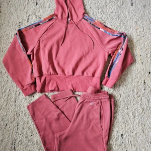 Korallfärgad hoodie och byxor från Adidas - Säljer ett set med en korallfärgad hoodie och matchande byxor från Adidas. Hoodien har en justerbar huva och dekorativa ränder på ärmarna. Byxorna har en elastisk midja och sidofickor. Perfekt för en avslappnad stil.Oanvänd!  Överdelen stl M Oversized! Byxa stl. S